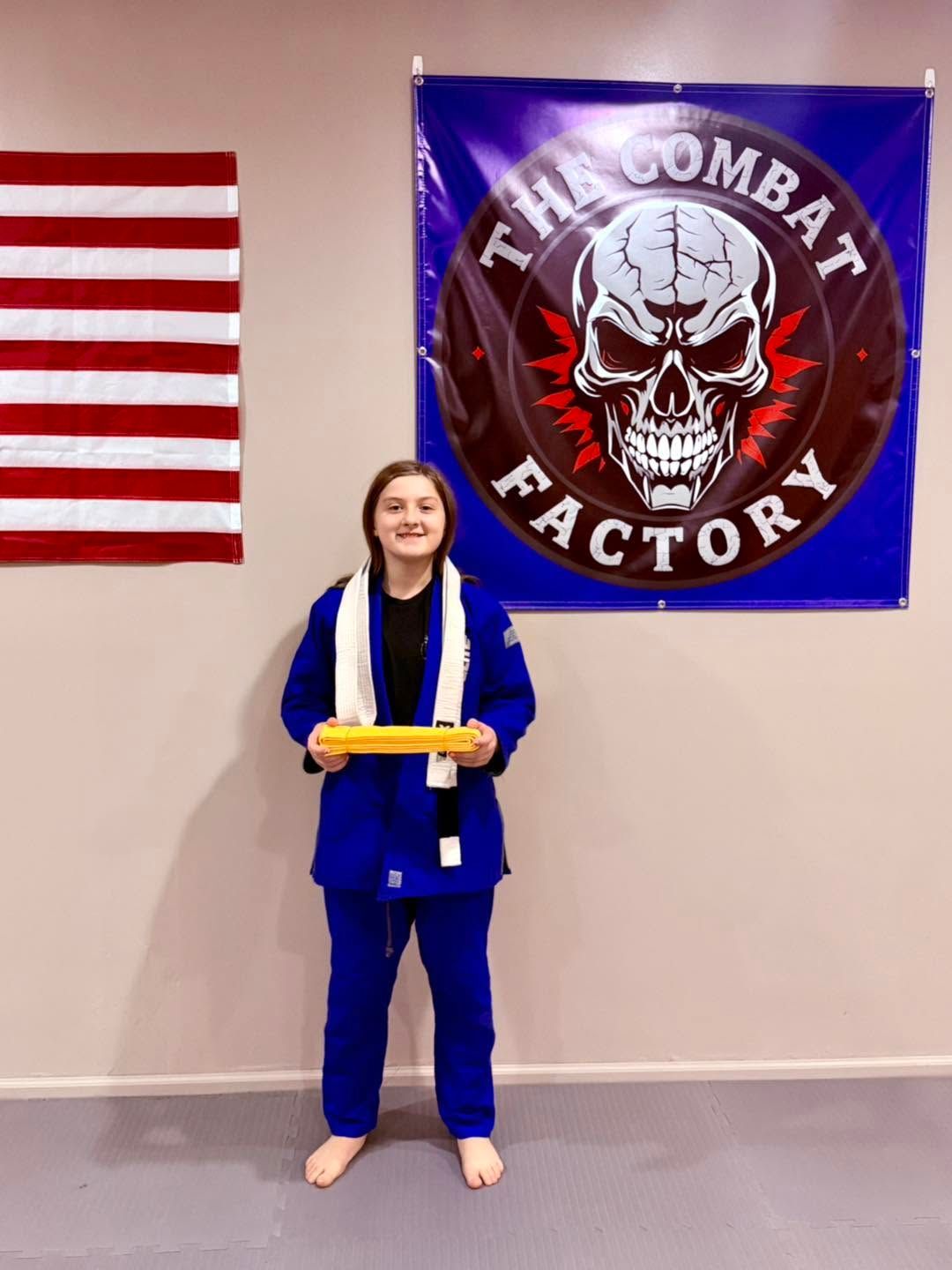 Person in blue martial arts uniform holding a yellow belt in front of an American flag and Combat Factory banner.