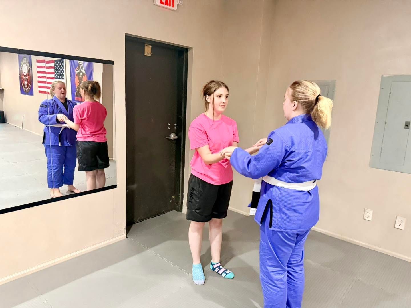Two women in blue uniforms practicing a handhold in a mirrored training room