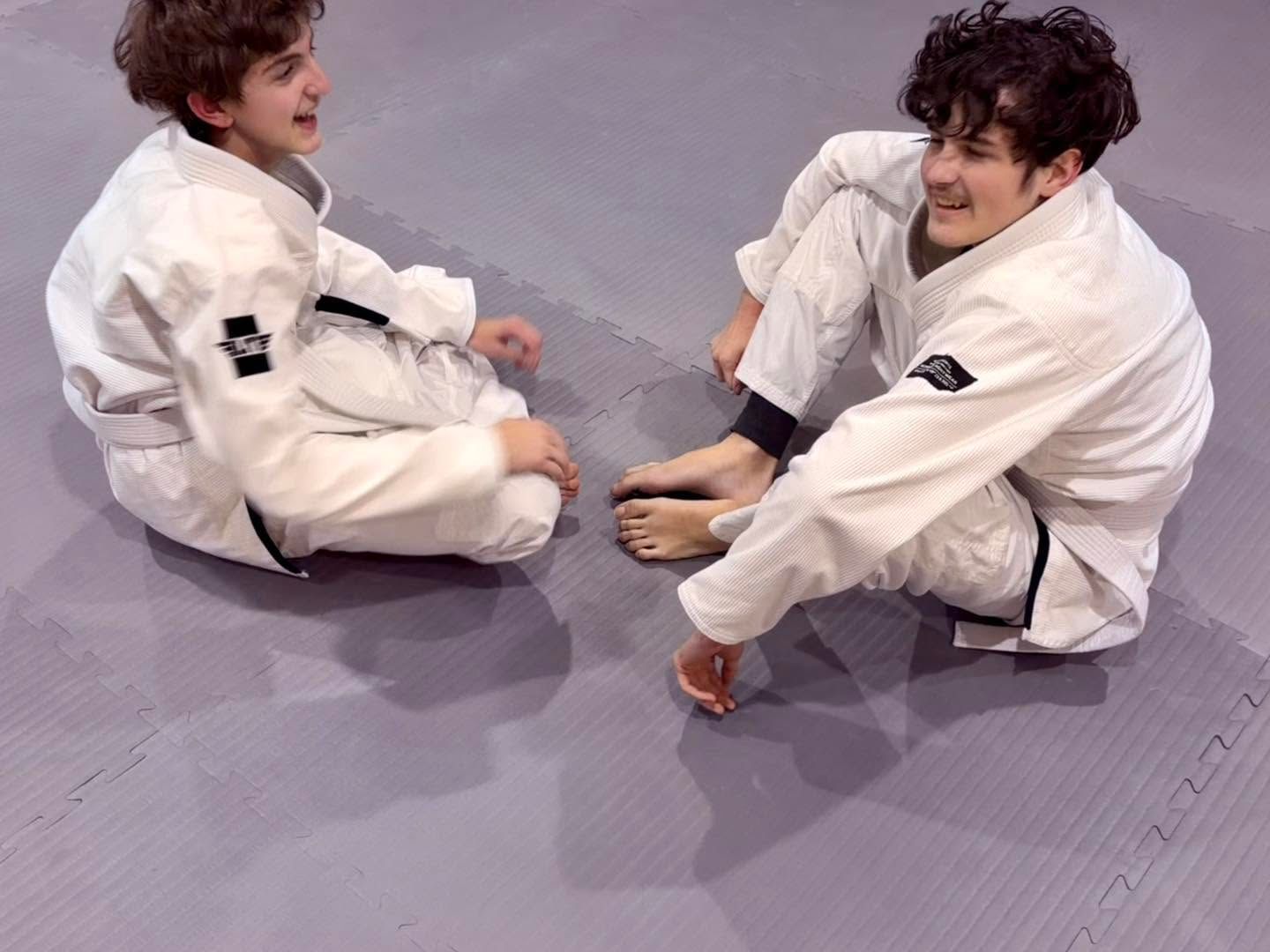 Two people in white martial arts gis sitting on a mat, smiling and holding each other’s ankles.