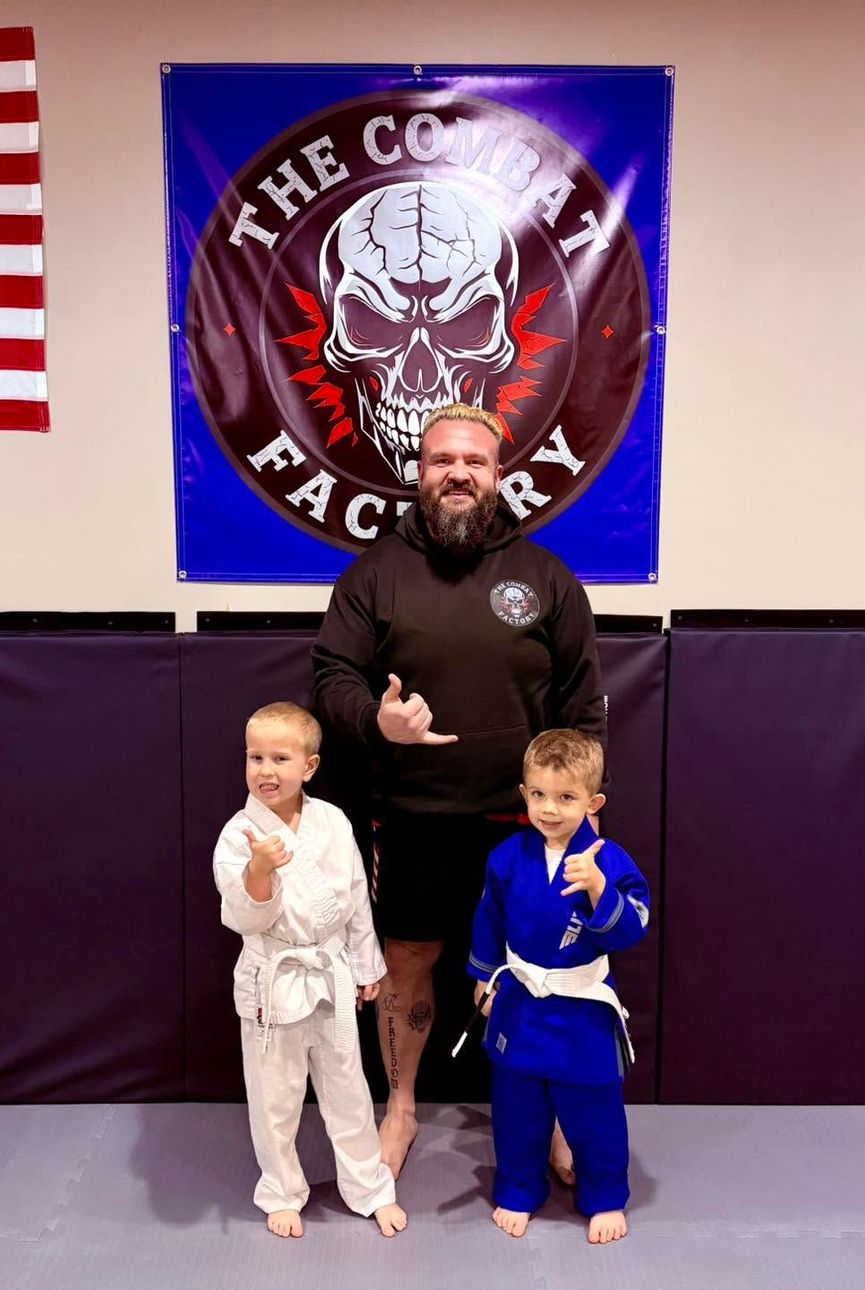 Three martial arts practitioners in gis pose in a dojo, with two children in front of a large skull banner.