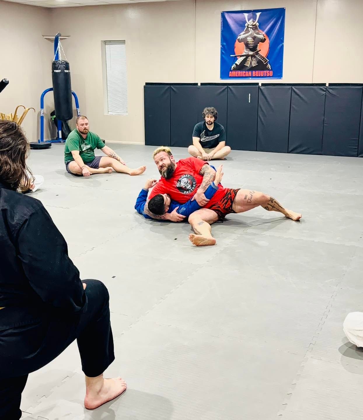 Two wrestlers in red and blue grapple on a gym mat while others watch from the sides.