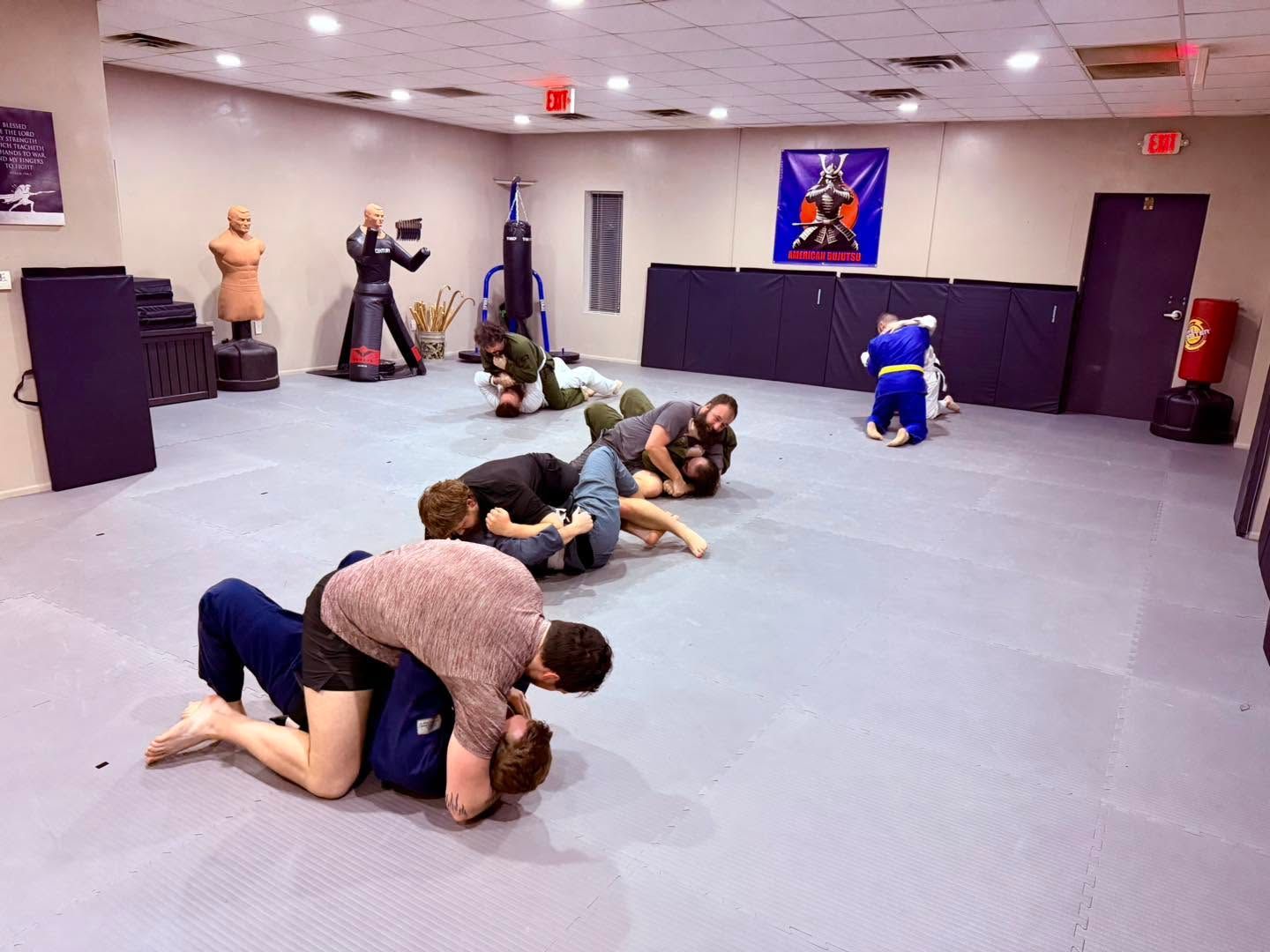 Jiu-jitsu class practicing grappling on mats in a bright training room with a wall poster and mirrors.