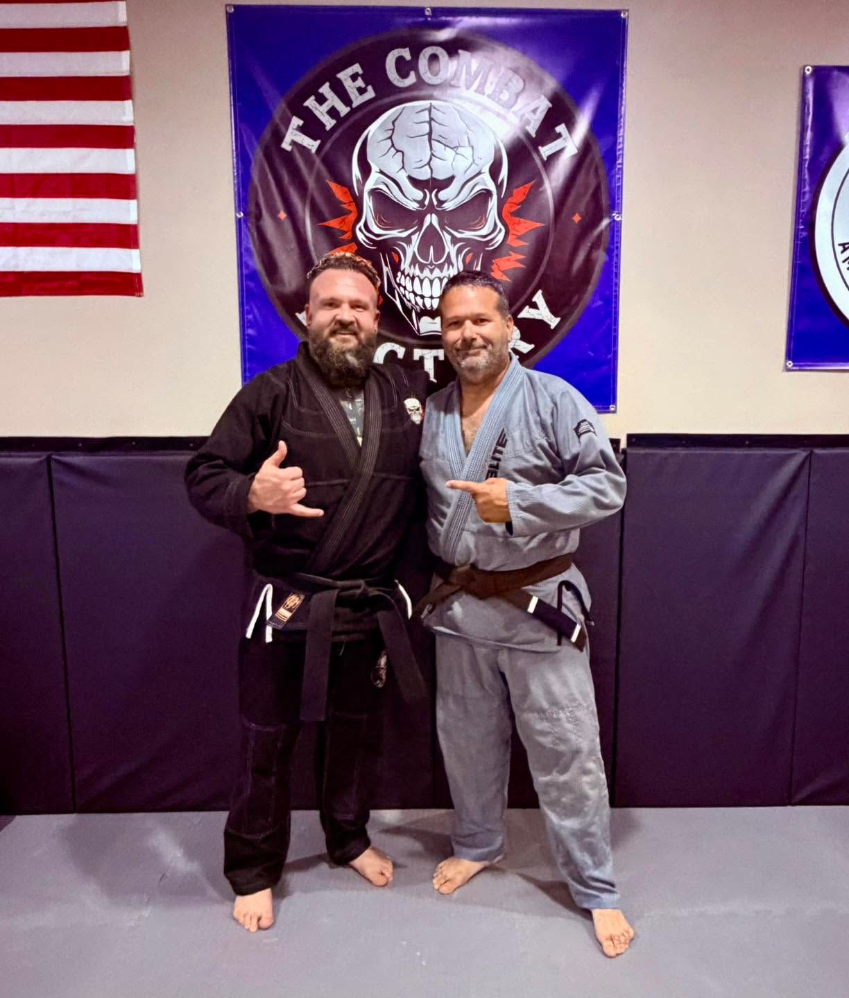Two men in martial arts gis pose on a mat in front of a skull banner, smiling and giving thumbs-up.