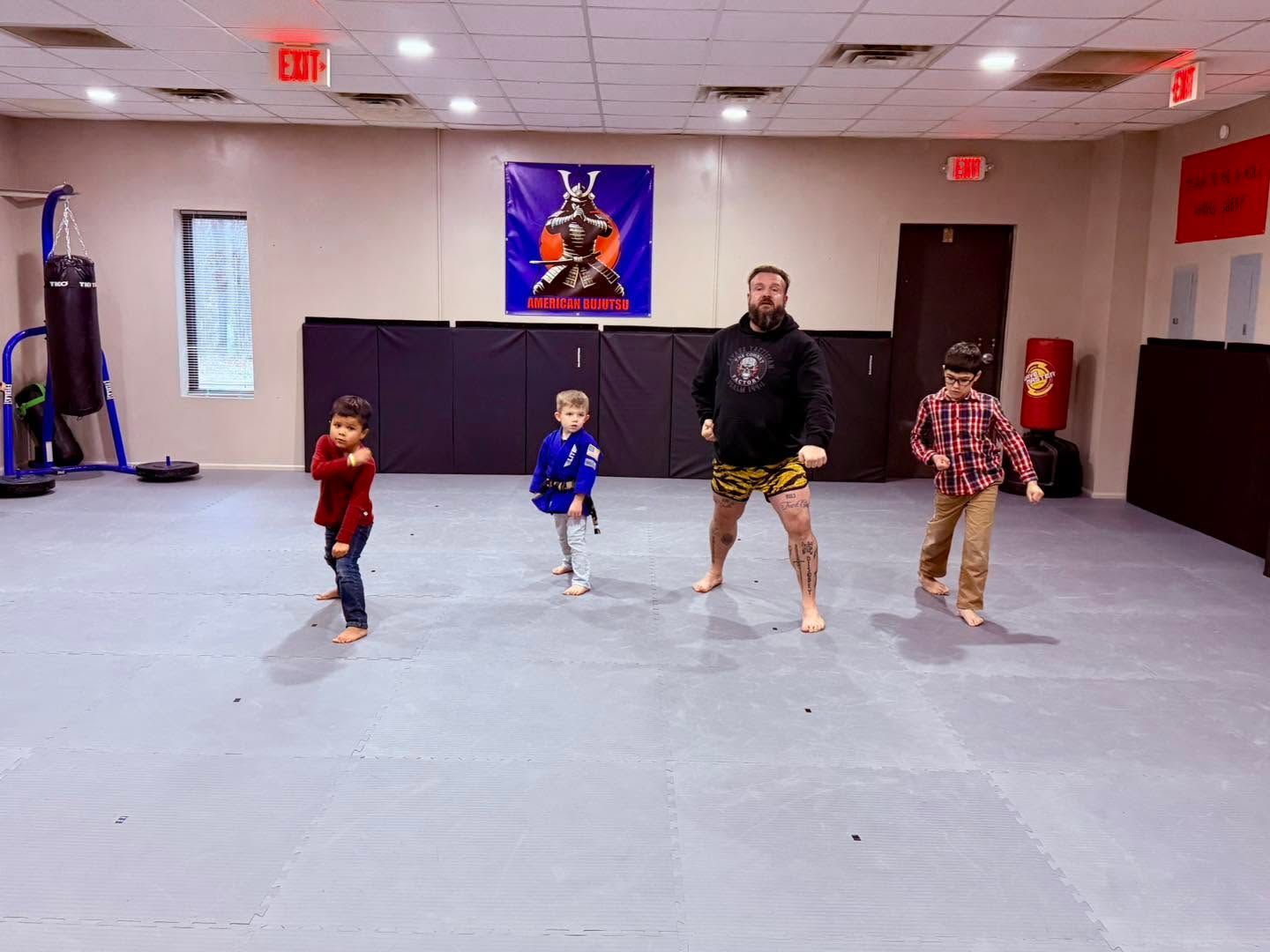 Four people posing in a dance studio, with a child in blue, child in red, adult in black, and child in plaid.