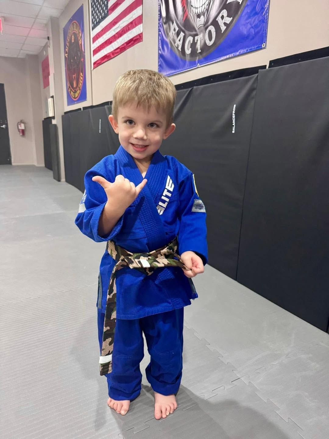 Young child in a blue martial arts gi with a black belt, standing and smiling in a dojo