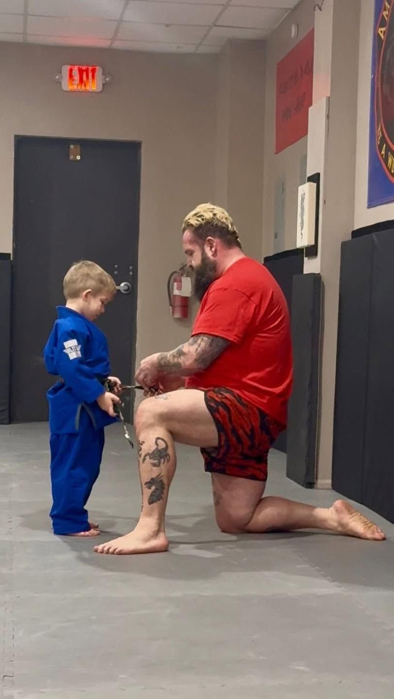 Adult kneeling with a child in a martial arts studio, both in blue and red training clothes.