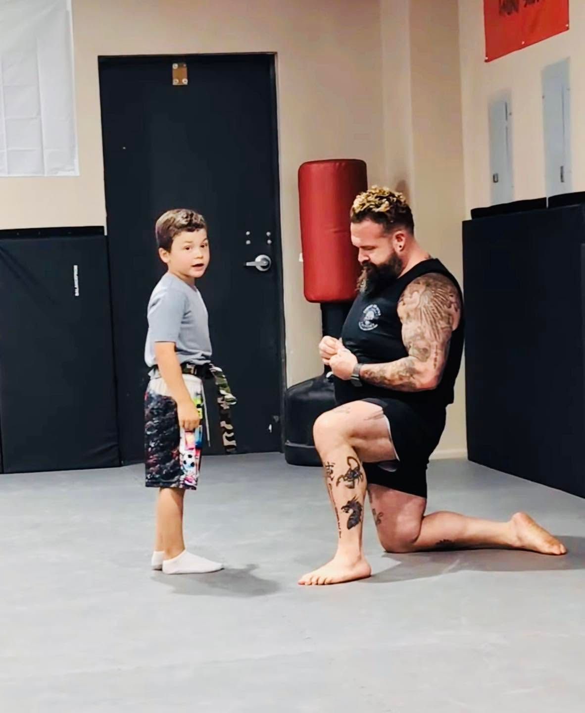 Man kneels beside a child in a gym, holding a training stance and looking toward the camera.