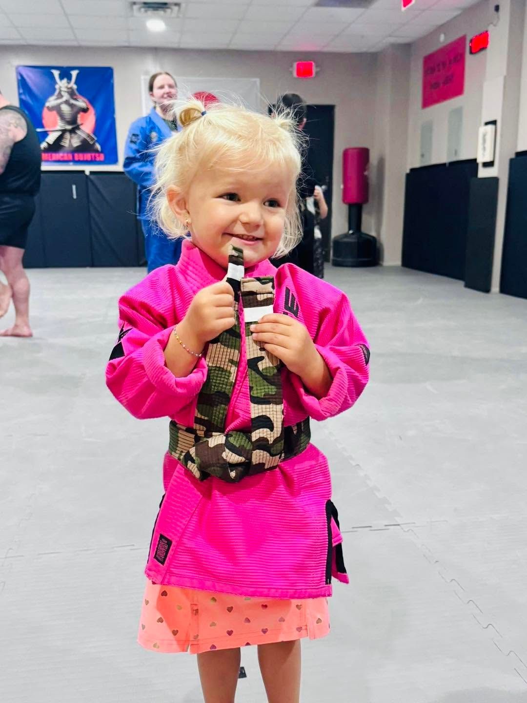 Smiling child in bright pink clothing holding a toy microphone in a gym or studio.
