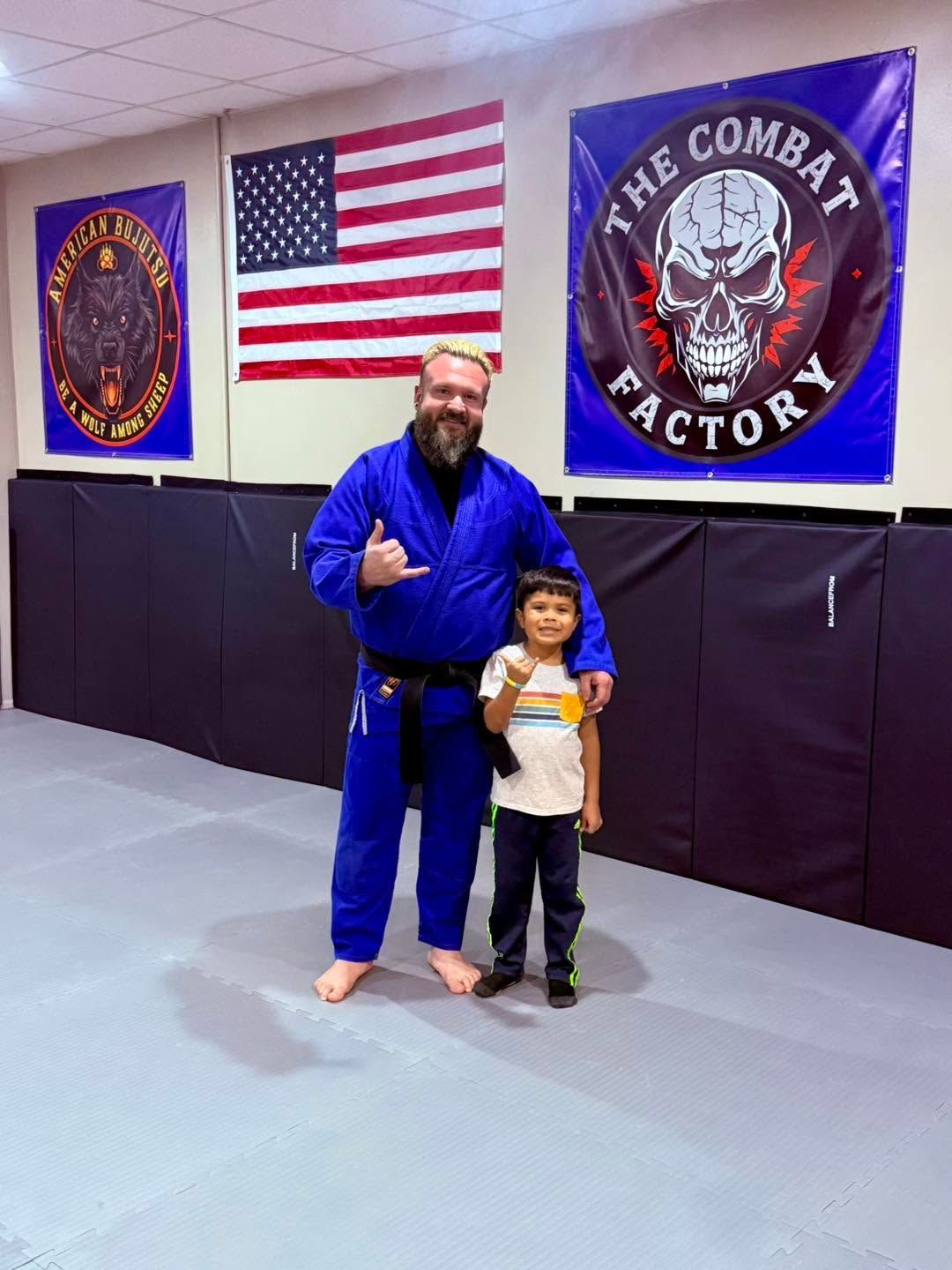 Two people in blue gis posing on a mat inside The Combat Factory gym, with flags on the wall.
