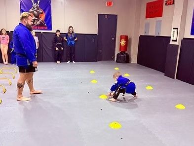 Child in blue crawls through yellow cones in a gym while an adult watches nearby.