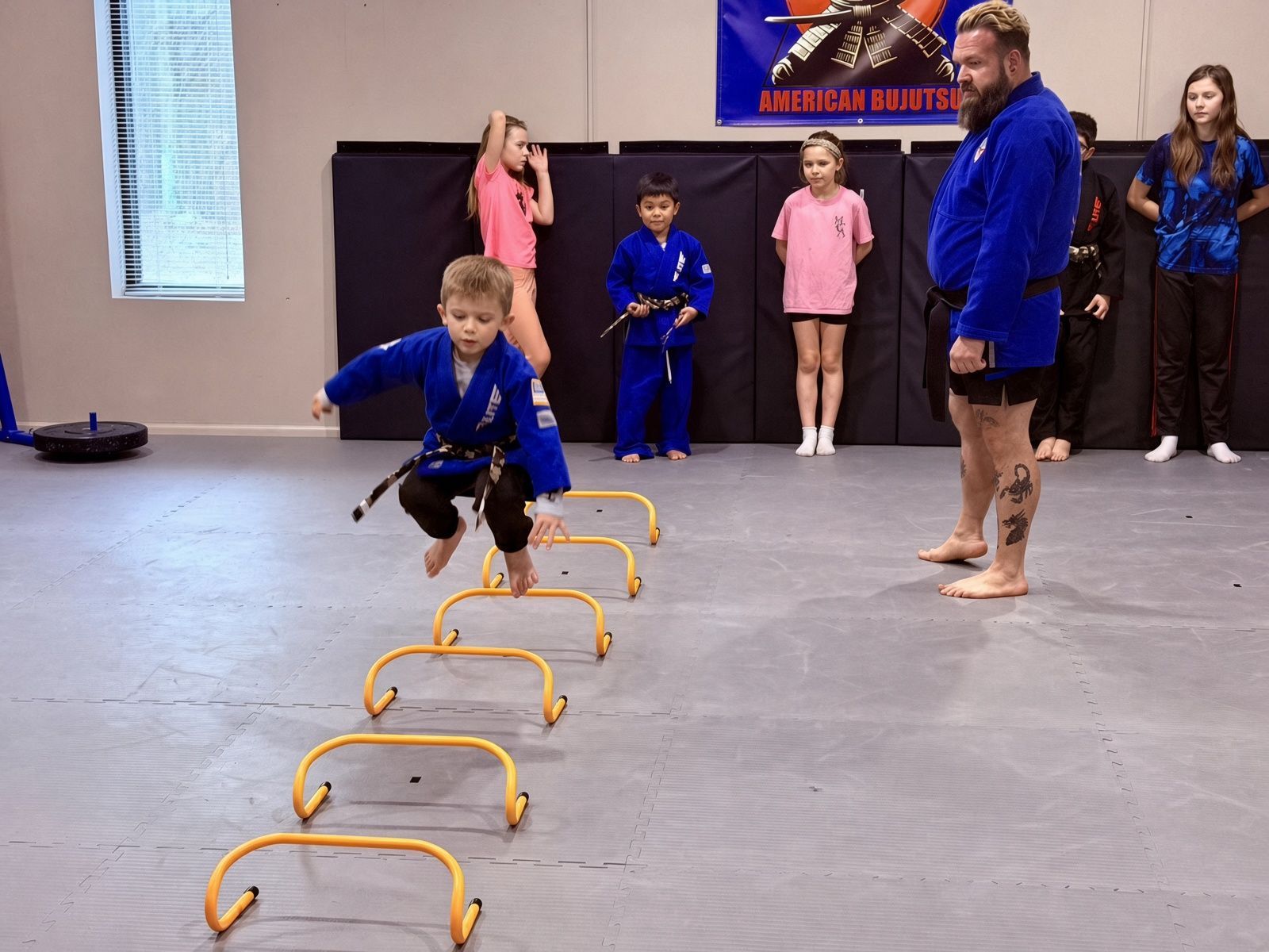 Child leaping over yellow hurdles in a gym while others watch from the side