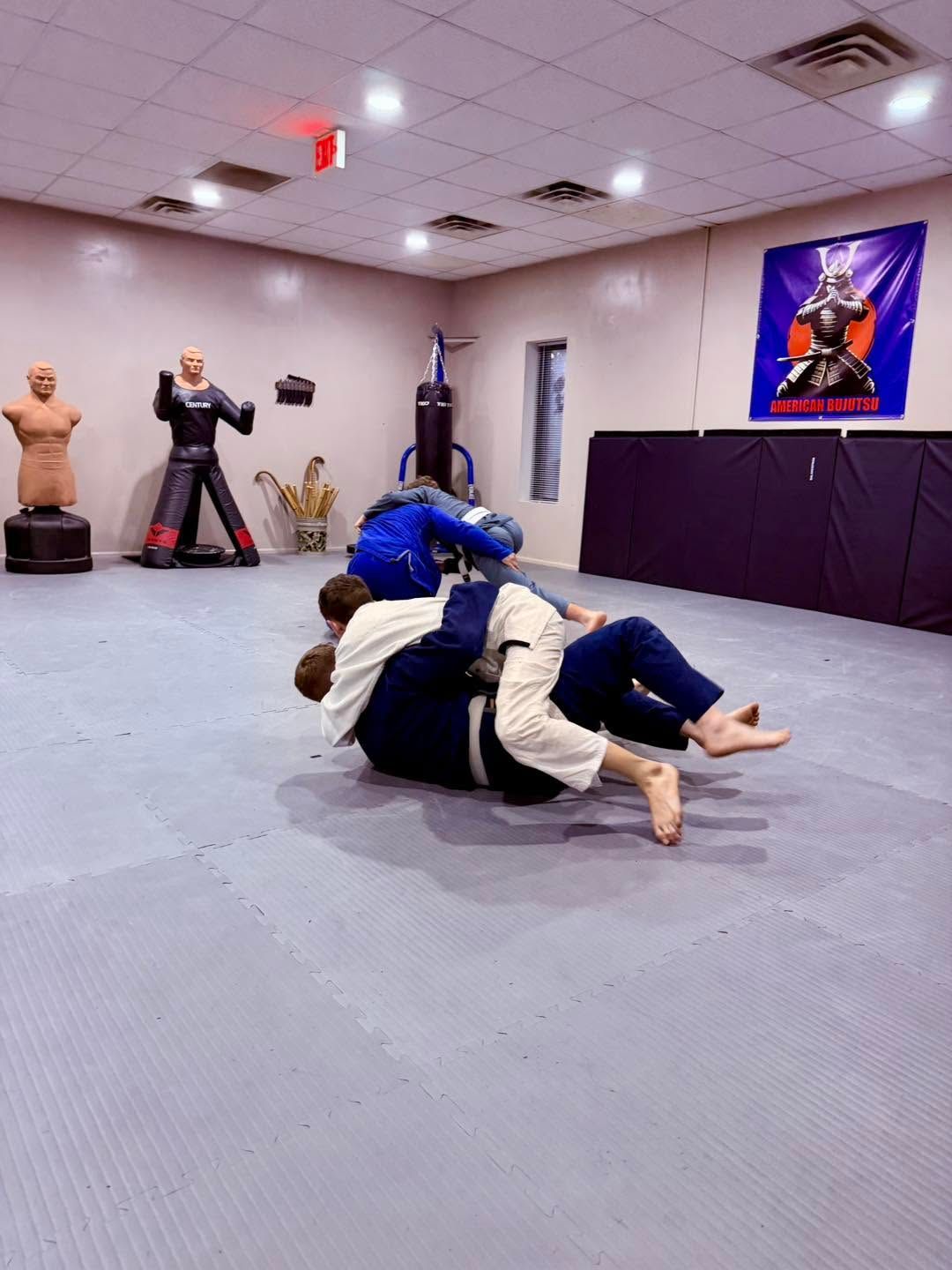 Martial arts class practicing grappling on mats in a dojo with training dummies and a wall poster