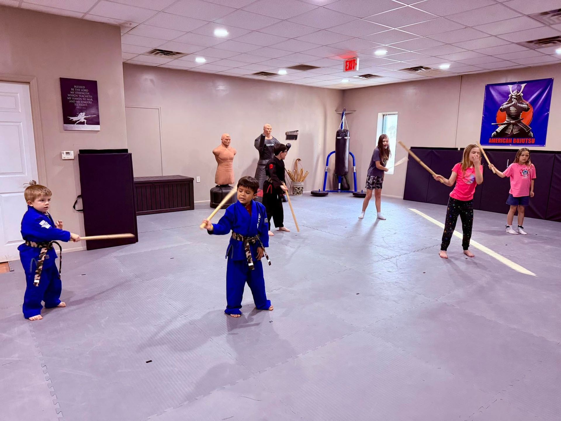 Children practicing martial arts in a studio, some with swords, facing an instructor near the back wall.
