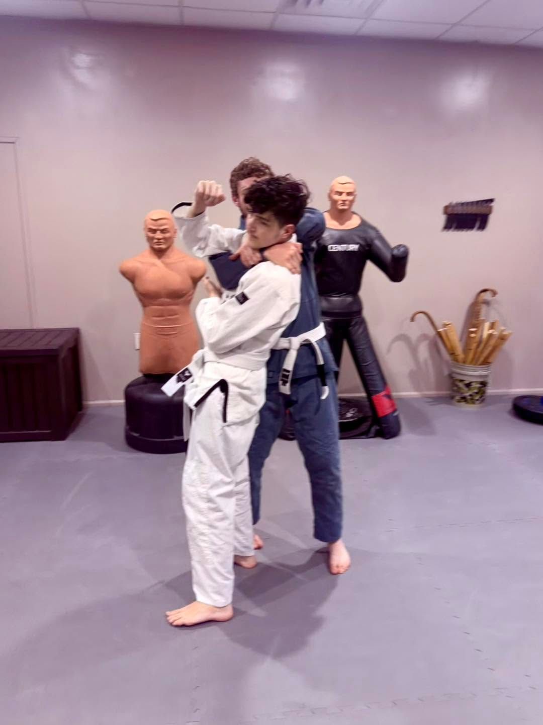 Two martial artists practicing a grappling move in a studio, with training dummies in the background.