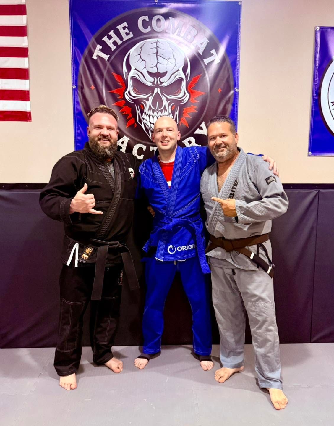 Three men in gi uniforms pose on a martial arts mat in front of a combat academy banner.
