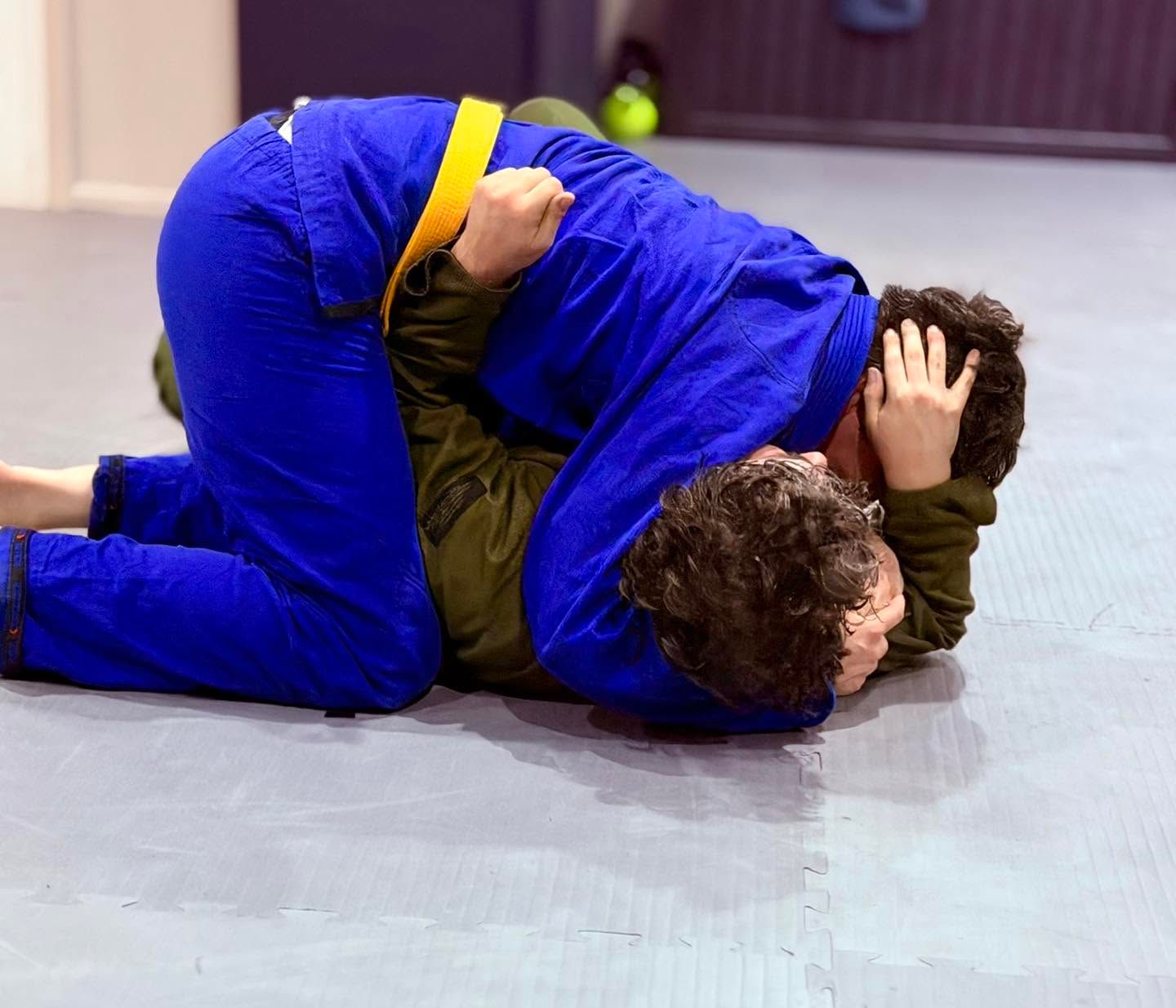 Two wrestlers in blue and green uniforms grappling on a mat during practice