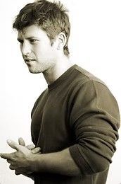 Man in a dark sweater standing in profile, looking left with hands clasped against a white background