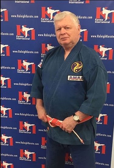 Man in a navy martial arts uniform holding a red belt in front of a branded step-and-repeat backdrop