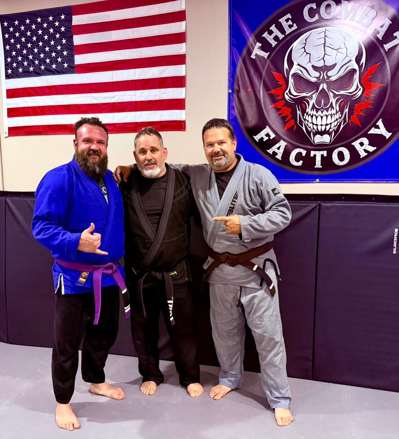 Three men posing barefoot in a gym, with an American flag and “The Combat Factory” banner behind them.