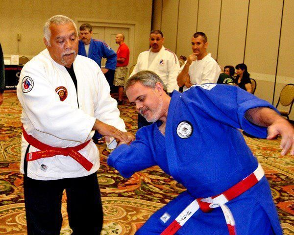 Two martial artists practicing a wrist lock in a hotel ballroom, one in a white gi and one in blue.