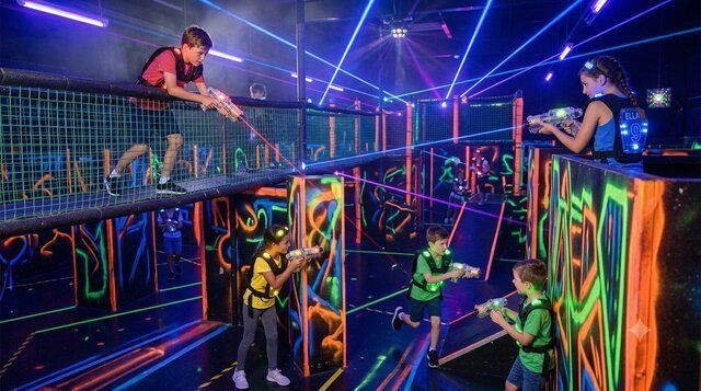 children in neon vests play laser tag in a dark arena with glowing walls elevated walkways and bright blue laser beams crisscrossing the room