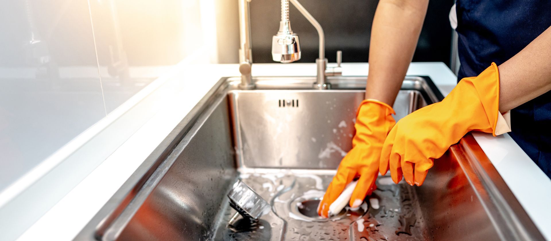 Person in orange gloves washing dishes in a stainless steel sink under running water.