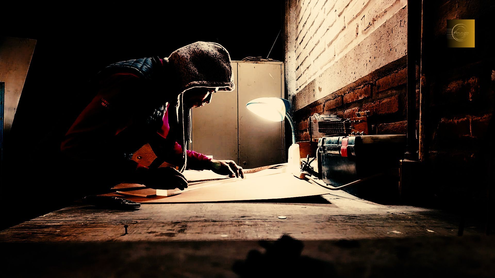 Una persona se agacha junto a una pared, trabajando a la luz de una lámpara de escritorio brillante en una habitación oscura y desordenada.