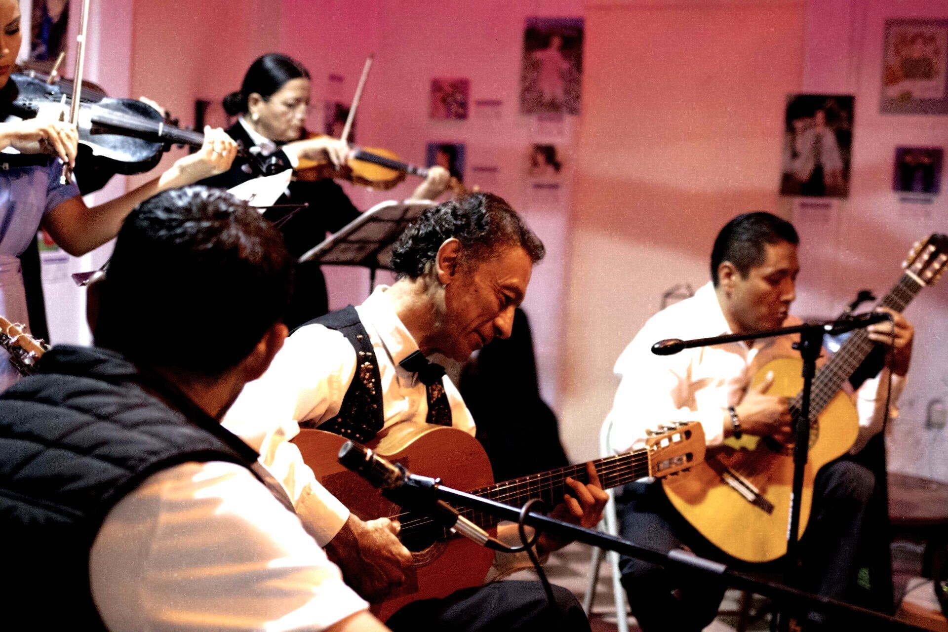 Hombre tocando la guitarra acústica en un taburete en una luminosa sala de música con piano y batería.