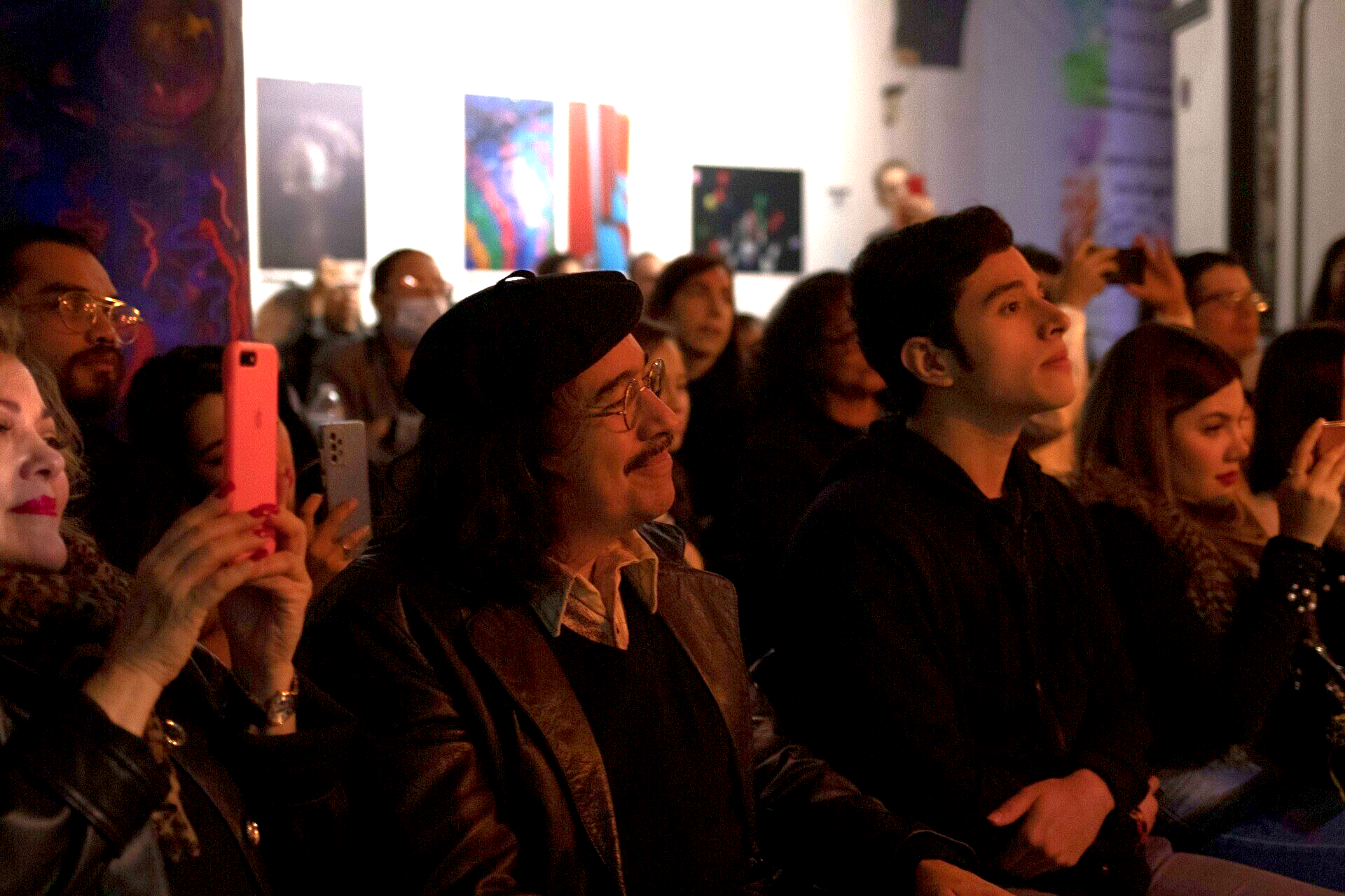 Público observando una presentación en una galería, rostros iluminados por una luz cálida, con coloridas obras de arte en la pared.