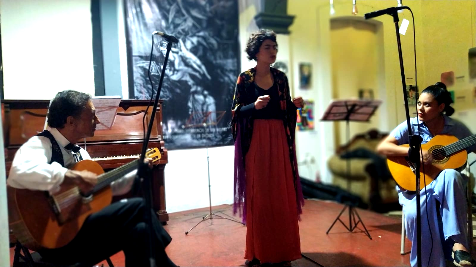 Tres músicos actúan en el interior: una cantante con un vestido rojo entre dos guitarristas sentados.