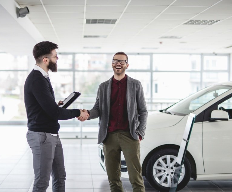 In un luminoso showroom, due uomini si stringono la mano accanto a un'auto bianca.