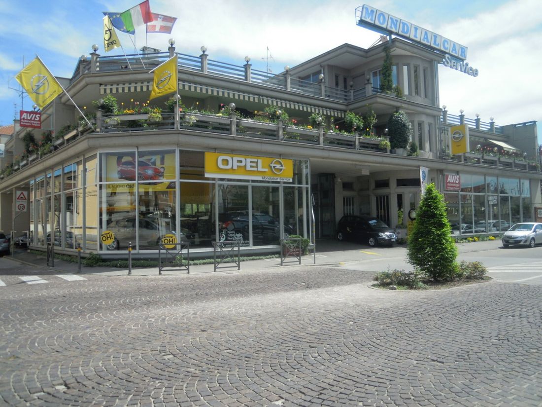 Edificio d'angolo con concessionaria Opel, bandiere sul tetto e auto parcheggiate su una strada acciottolata.