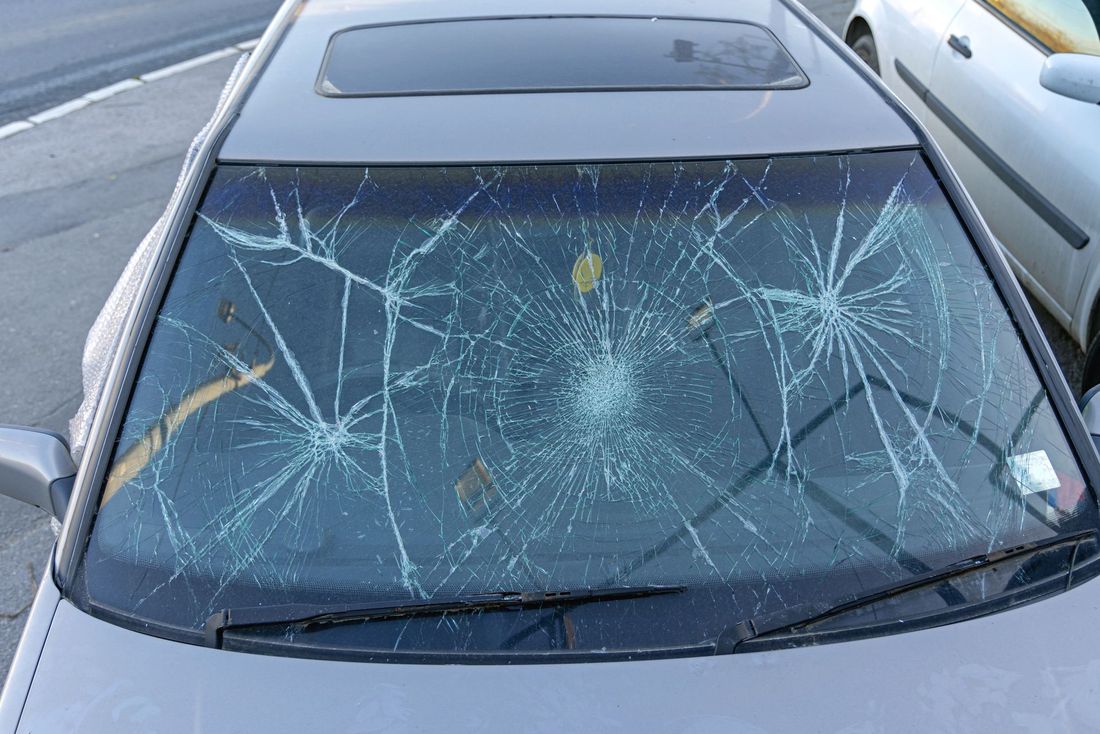 Parabrezza dell'auto in frantumi con ampie crepe e un punto d'impatto visibile, parcheggiata accanto a un'altra auto