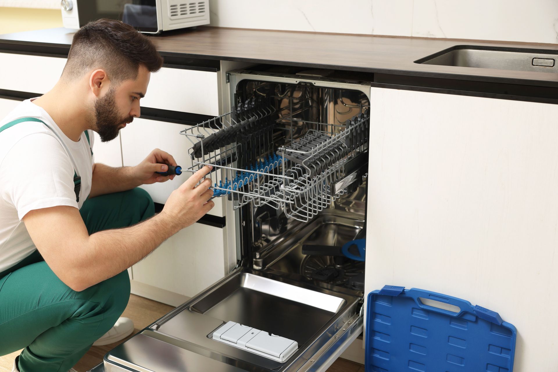 Technician inspecting the open door of a built-in dishwasher in a kitchen, with tools nearby.