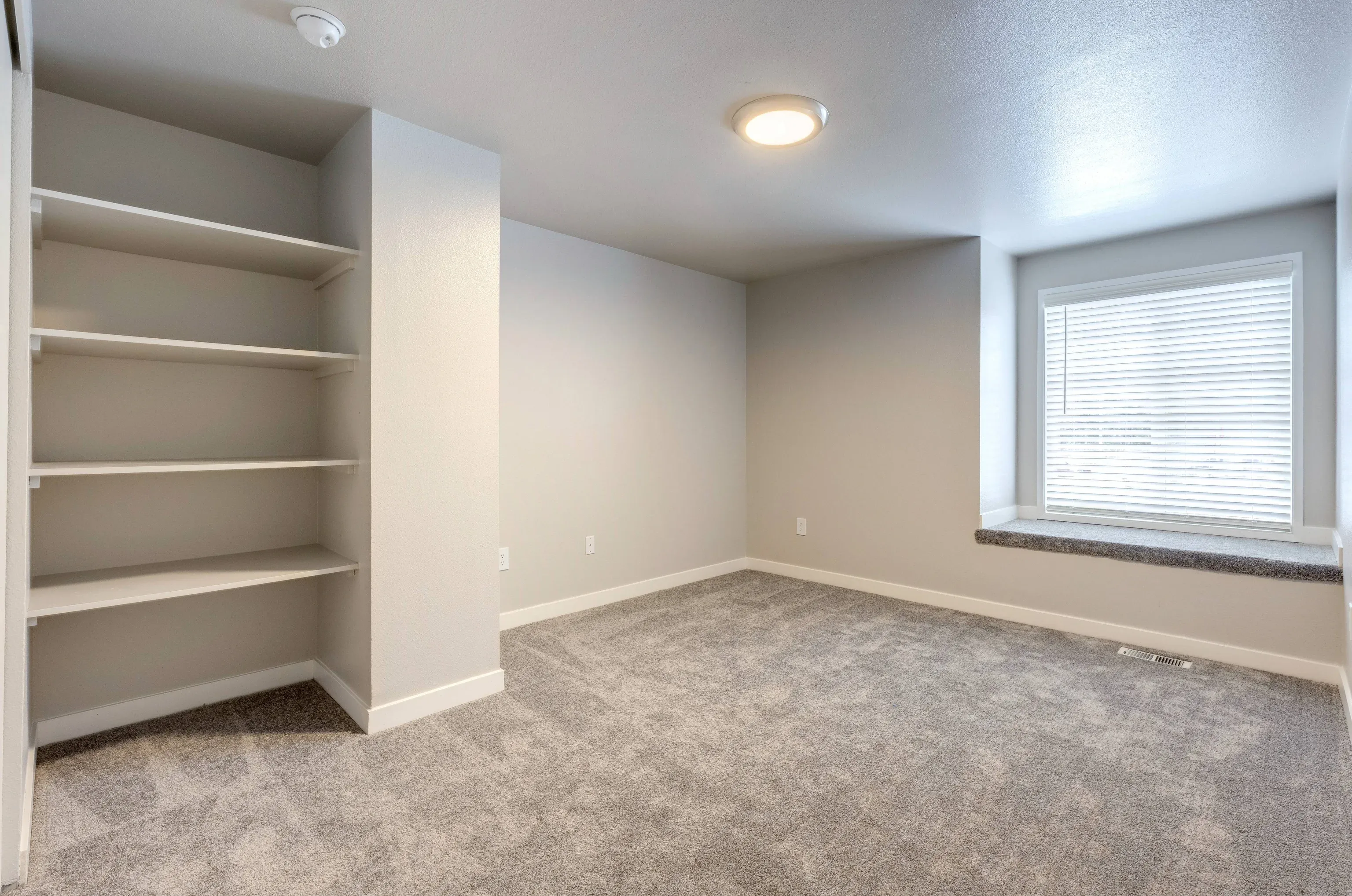 Carpeted room with built-in shelves on the left and a window with blinds on the right.