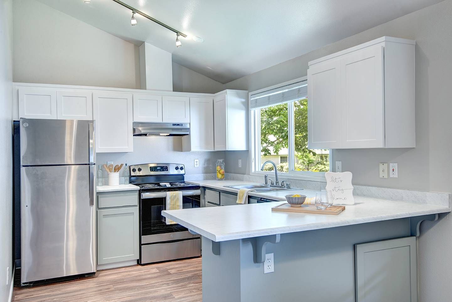 Bright apartment kitchen with white cabinets, stainless fridge and stove, and a window over the sink.