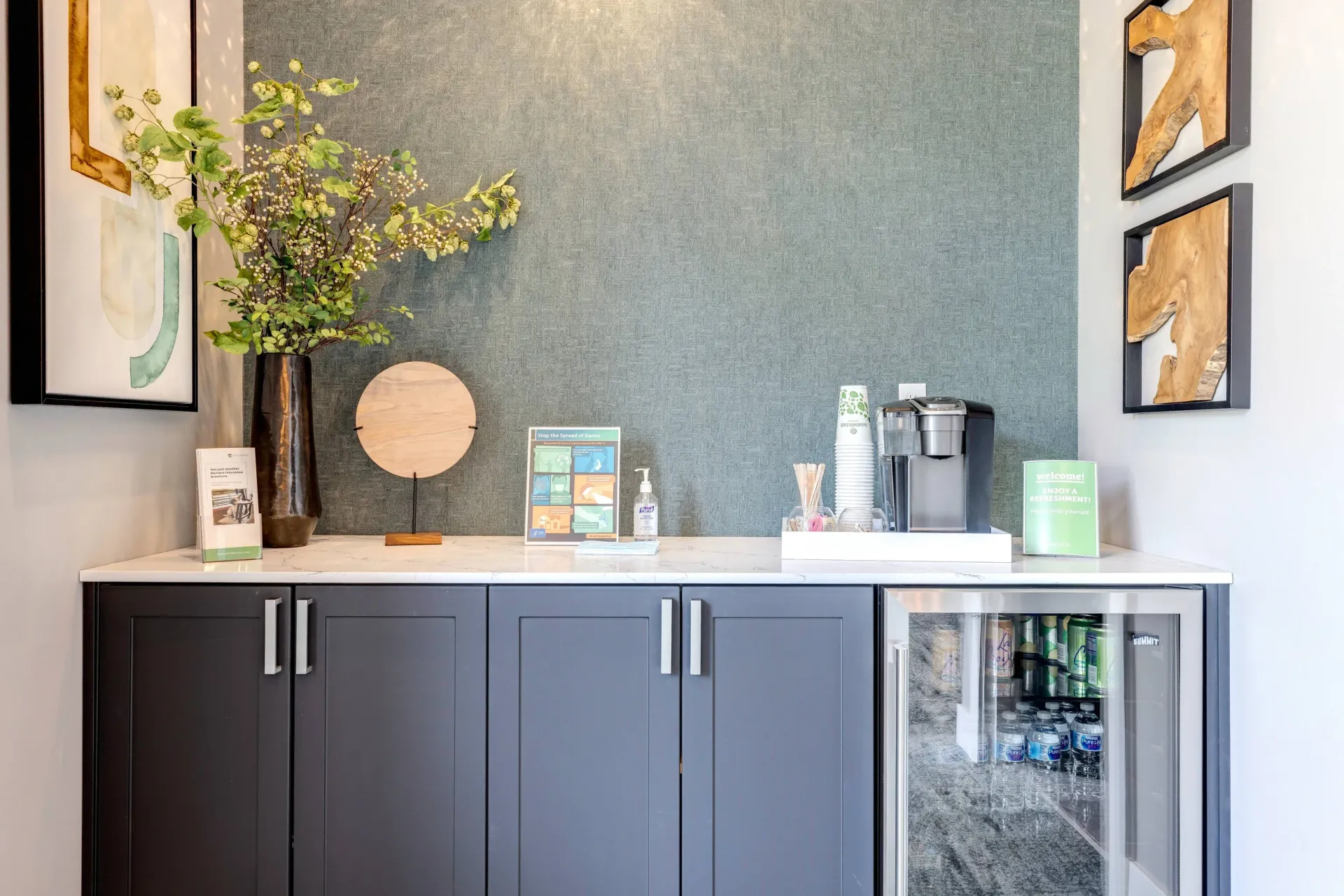 Community coffee bar with dark cabinets, a beverage fridge, and a coffee station against a blue textured wall.