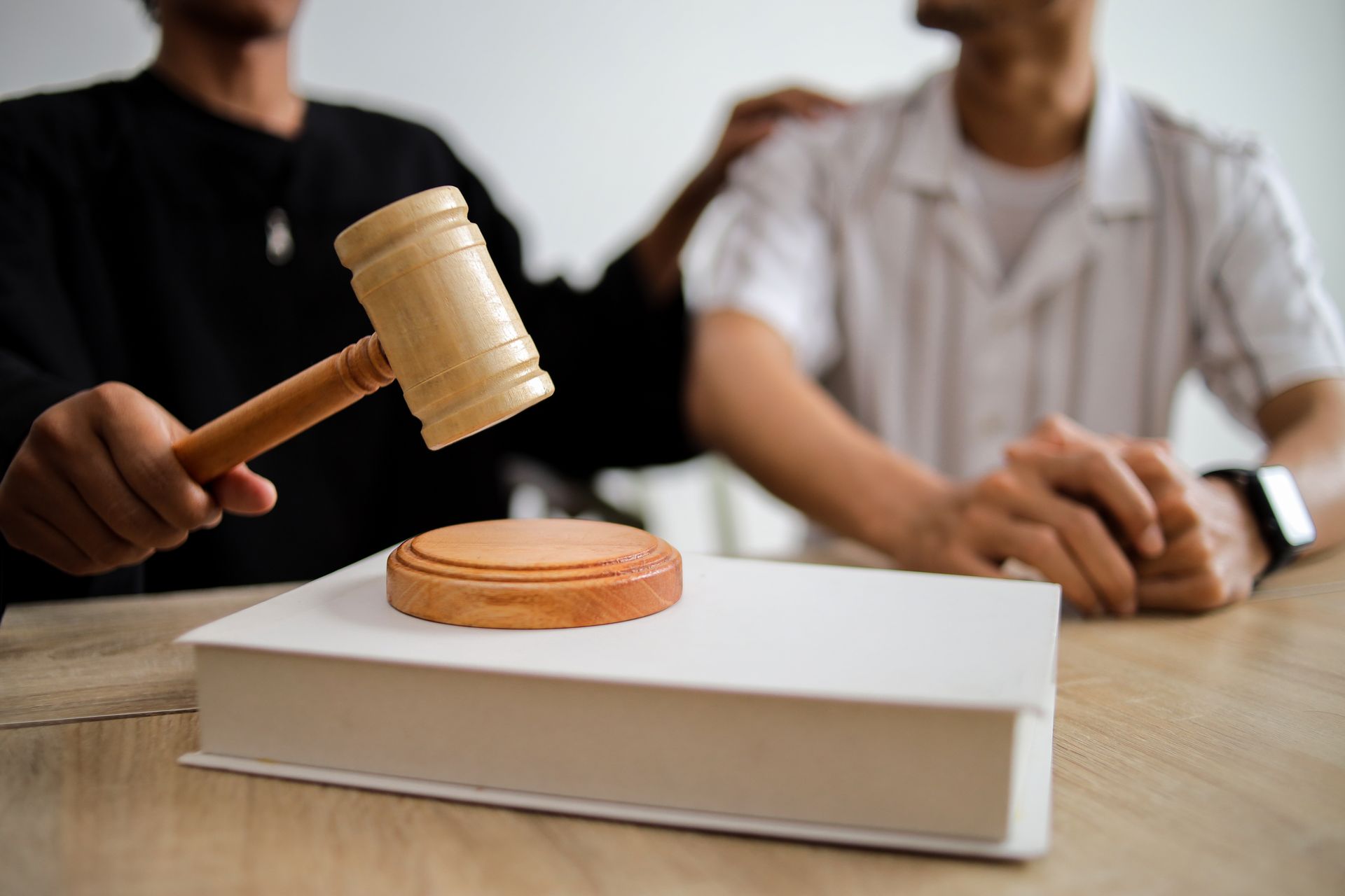 A lawyer sits next to a client holding a wooden hammer over a white-covered book.