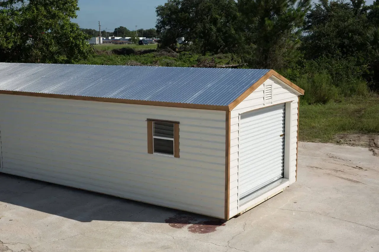 White storage shed with a corrugated metal roof, a roll-up garage door, and a small window.