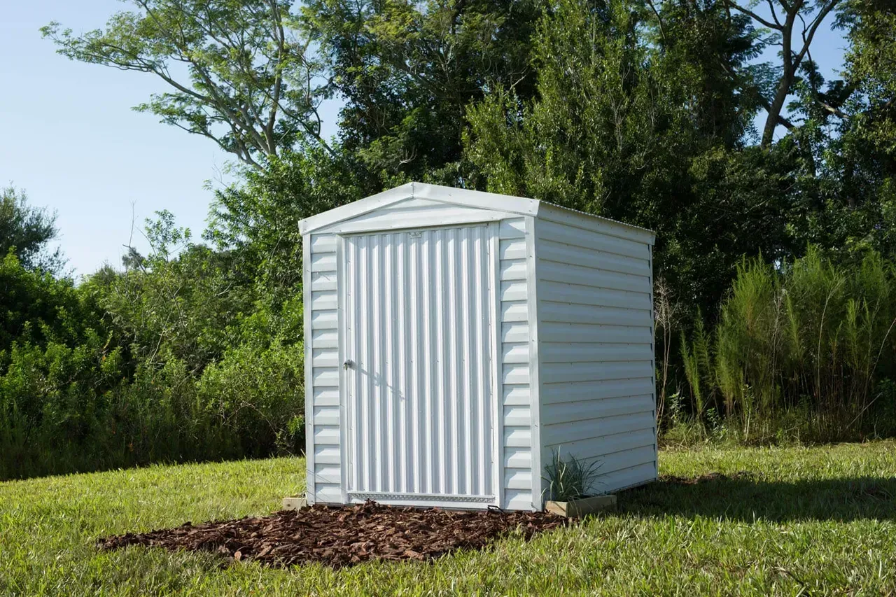 White metal storage shed in a grassy yard, surrounded by trees under a blue sky.