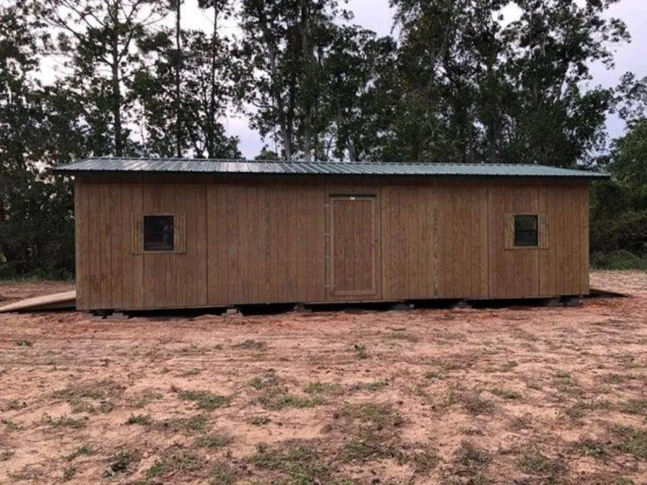 Wooden shed with a dark green roof, door, and two windows in a dirt field with trees.