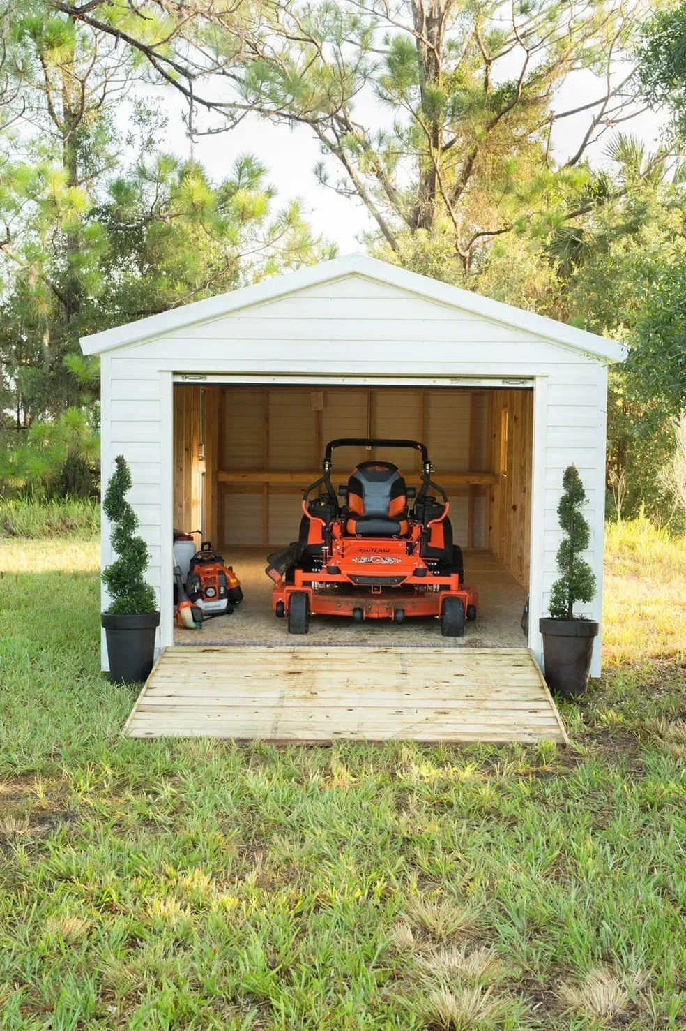 White shed with open doors, housing a riding mower and lawn mower, on grass.