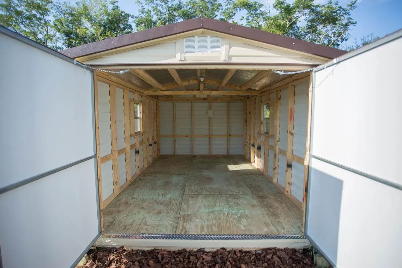 Interior view of an empty shed with open doors, wood framing, brown roof, and small windows.