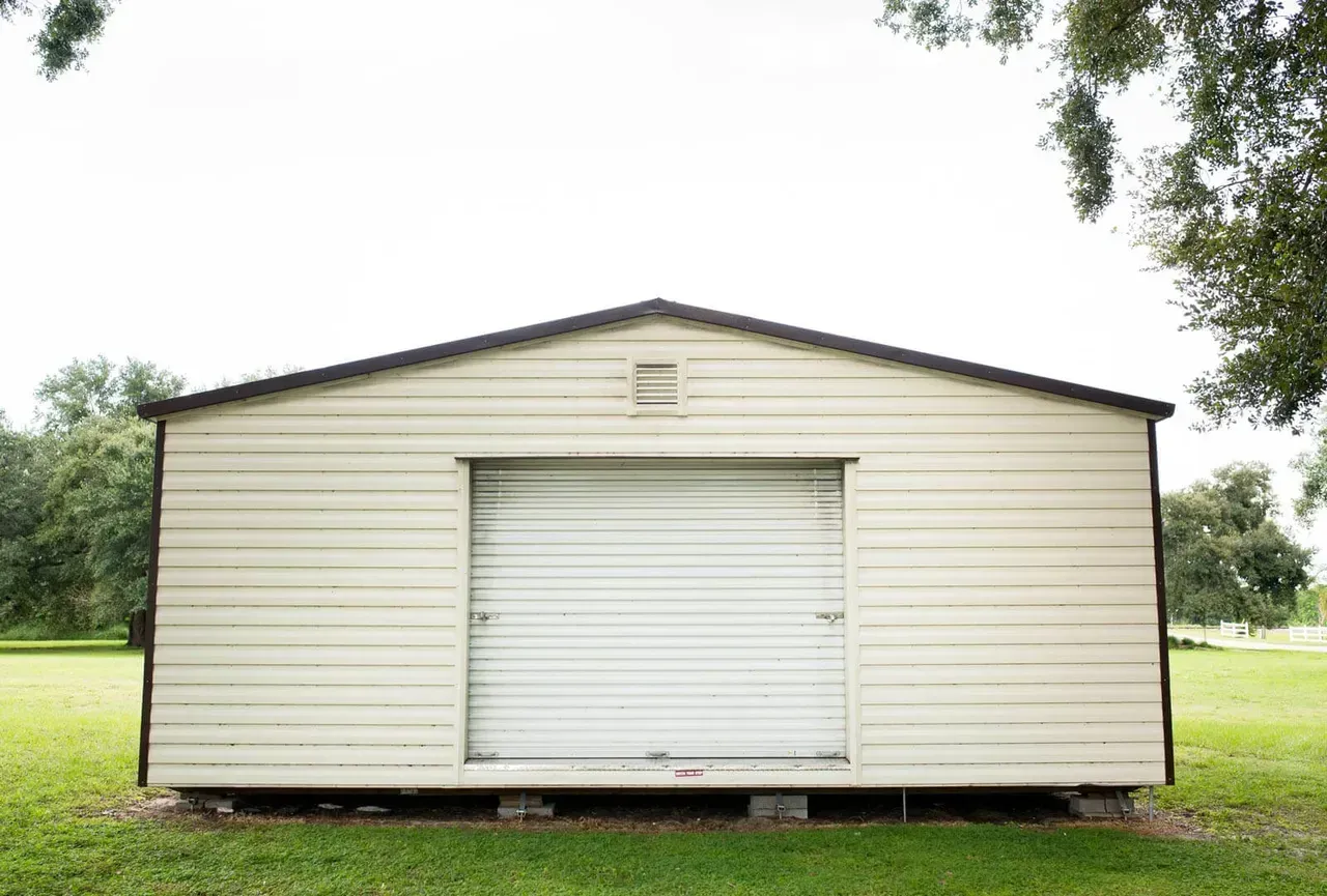 White shed with a roll-up door, set on concrete blocks. Green grass and trees in the background.