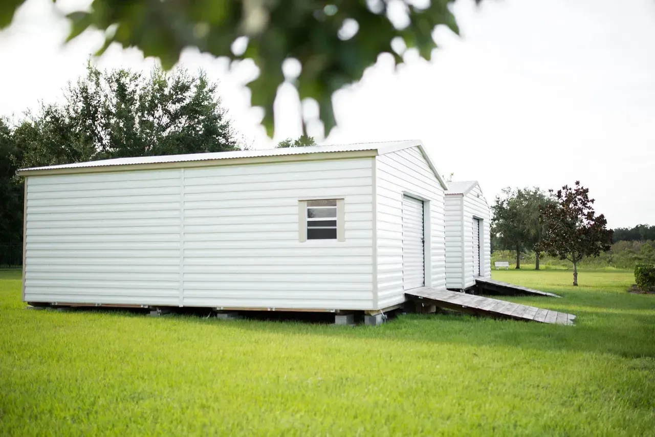 White sheds with ramps on a grassy lawn, trees in the background, overcast sky.