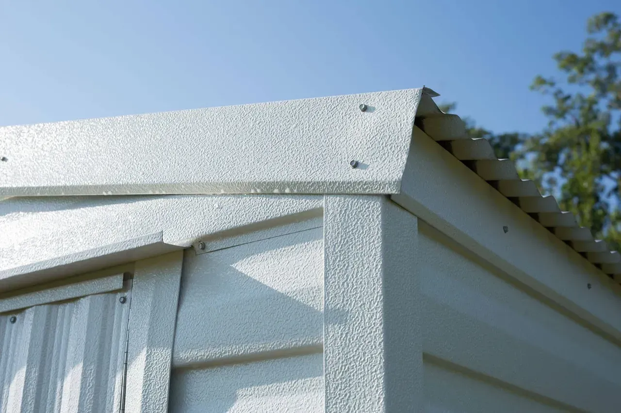 Close-up of a shed corner. The roof and walls are textured metal, light gray in color, against a blue sky.