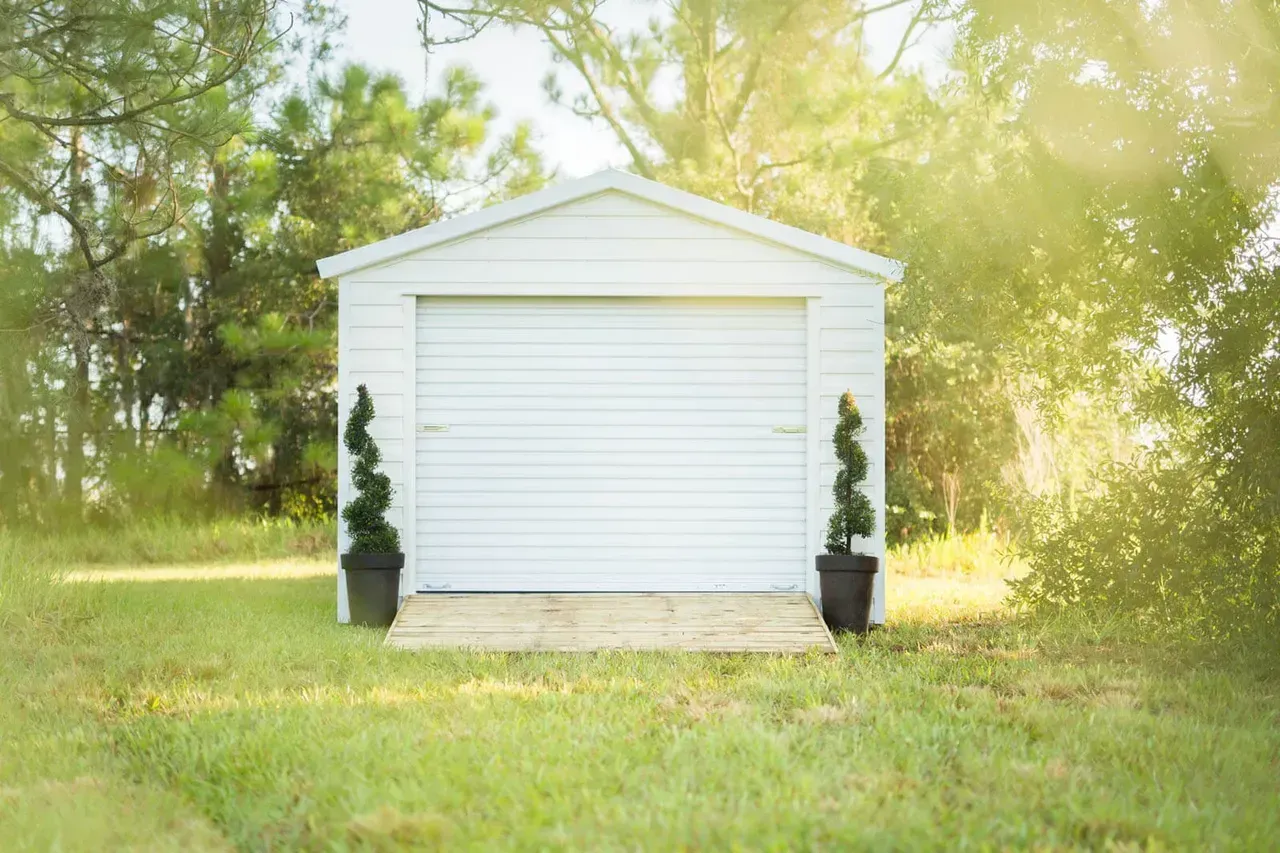 White shed with closed garage door in a grassy yard, flanked by potted topiaries, sunny background.