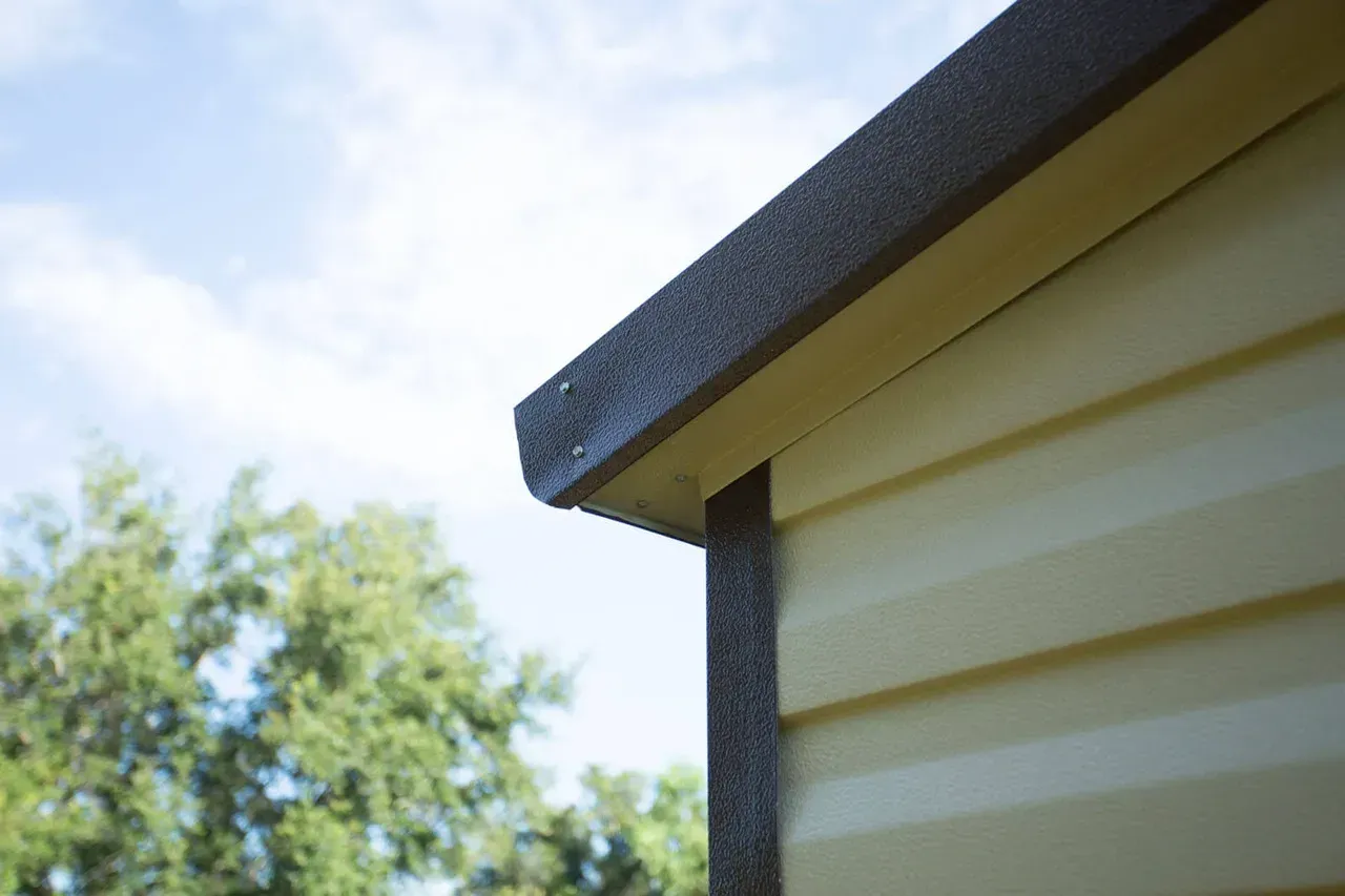 Close-up of a house corner with light yellow siding and brown trim under a blue sky.