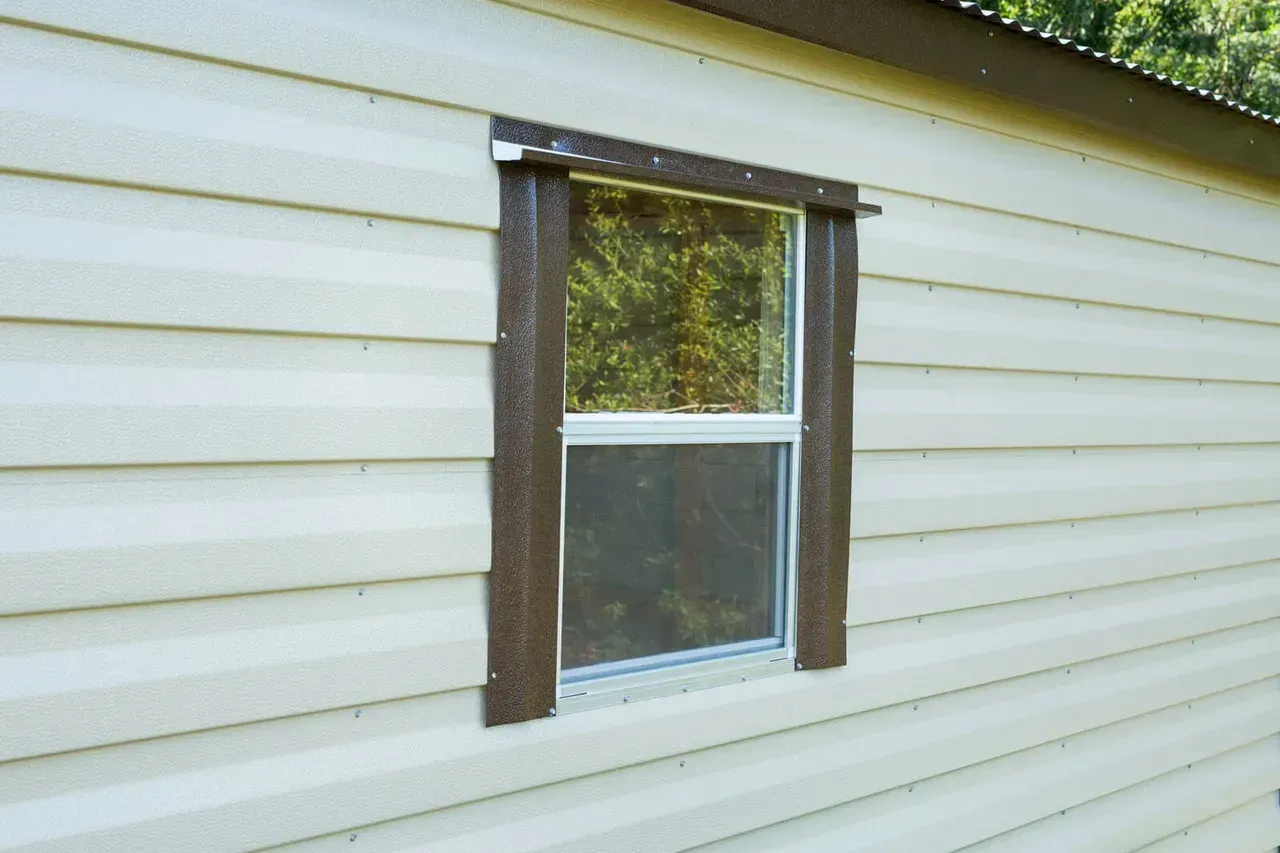 Window with brown trim on a beige siding wall, reflecting trees.