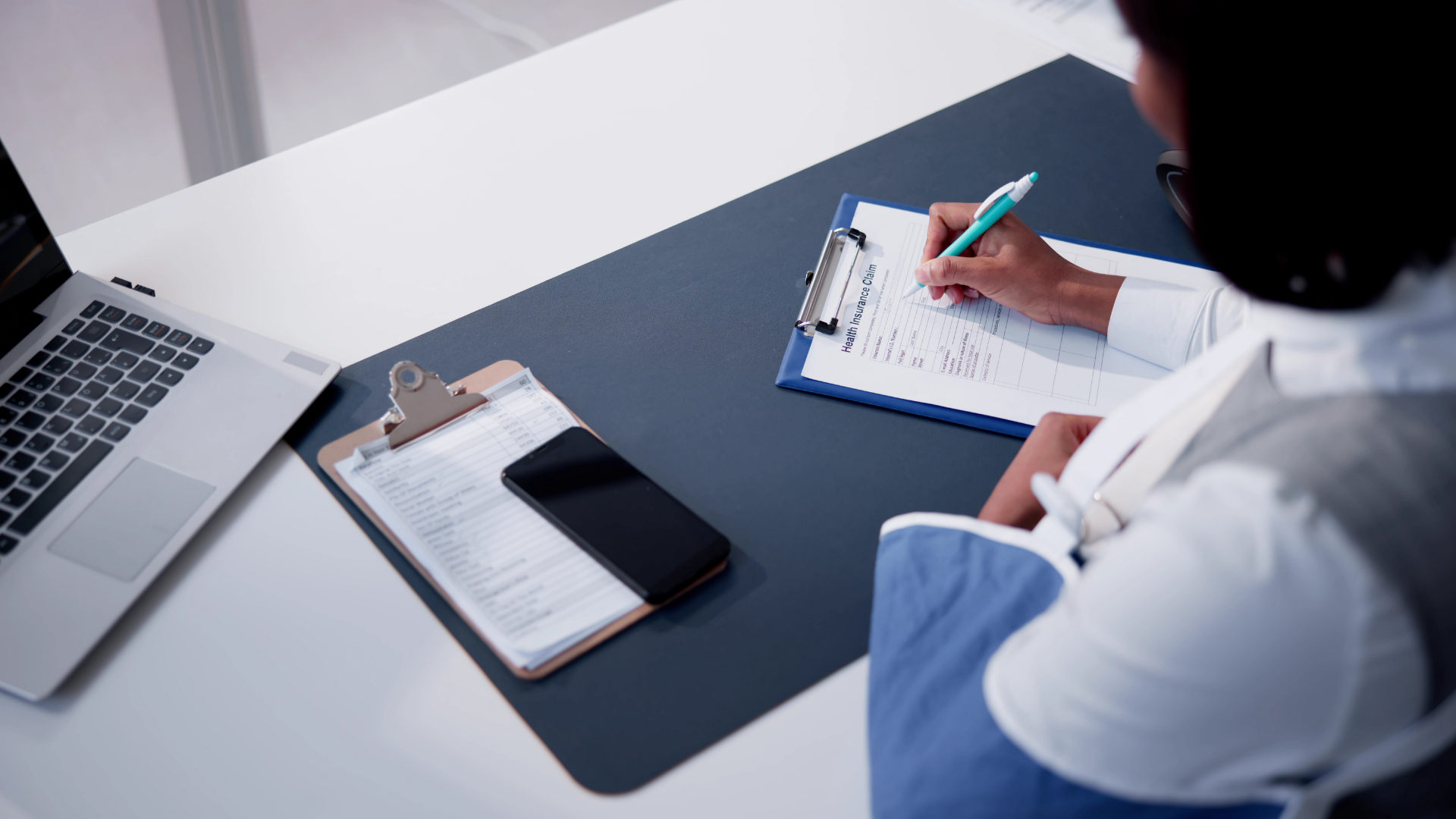 Person writing in a notebook at a desk with a laptop and clipboard, in a bright office setting